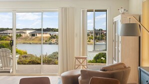 A room with a large sliding glass door leading to a deck with a view of a lake and houses. There is a brown leather sofa, a wooden chair, and a small wooden stool. A tall floor lamp is on the right side of the room.