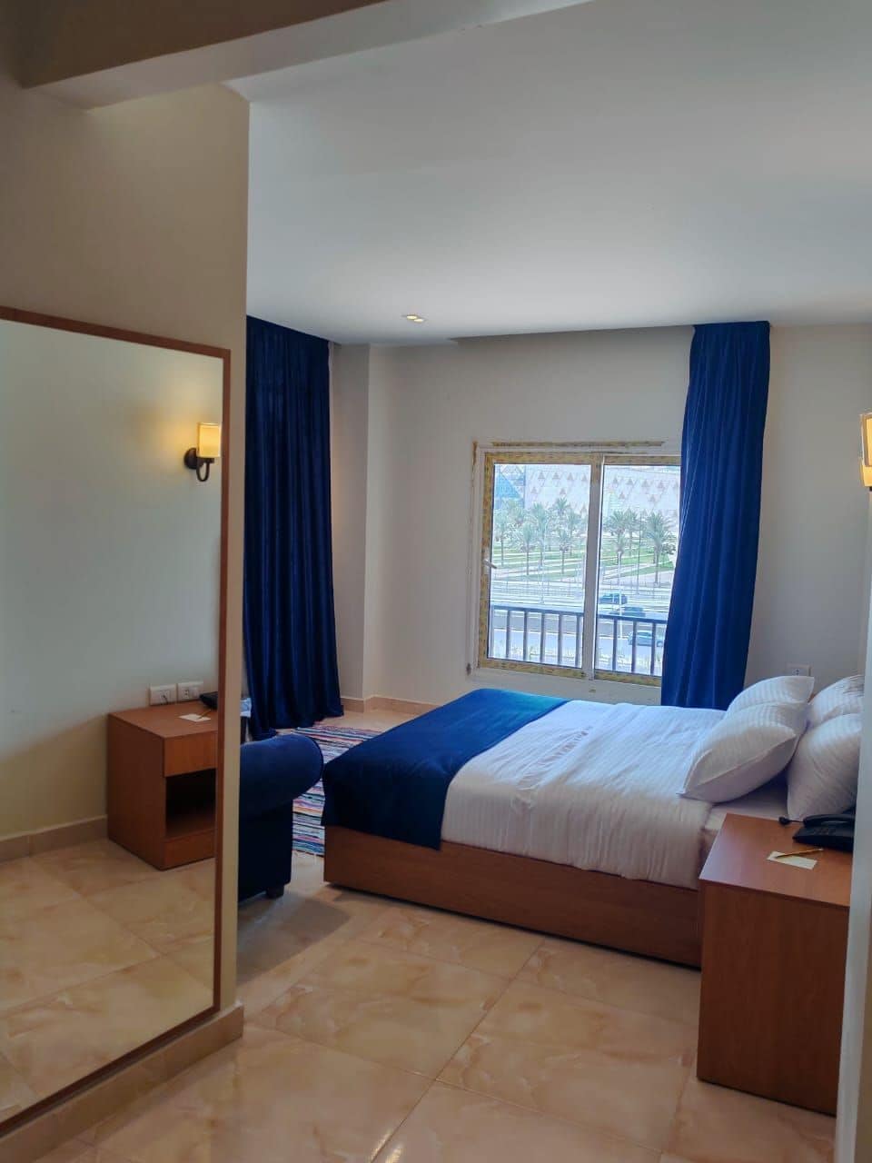 A hotel room with a bed, a nightstand, a mirror, a window with curtains, and a view of the outside.