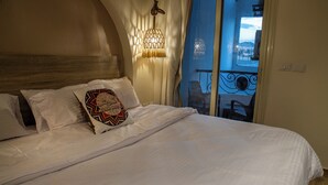 Una cama bien hecha con sábanas blancas y un cojín estampado, una lámpara colgante y vistas a un balcón exterior a través de una ventana.