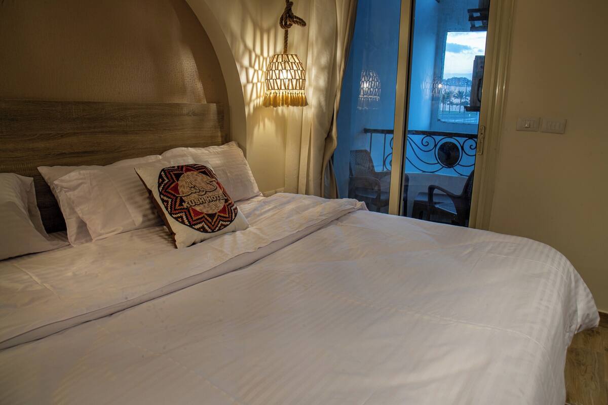 A neatly made bed with white linens and a patterned pillow, a hanging light fixture, and a view of an outdoor balcony through a window.