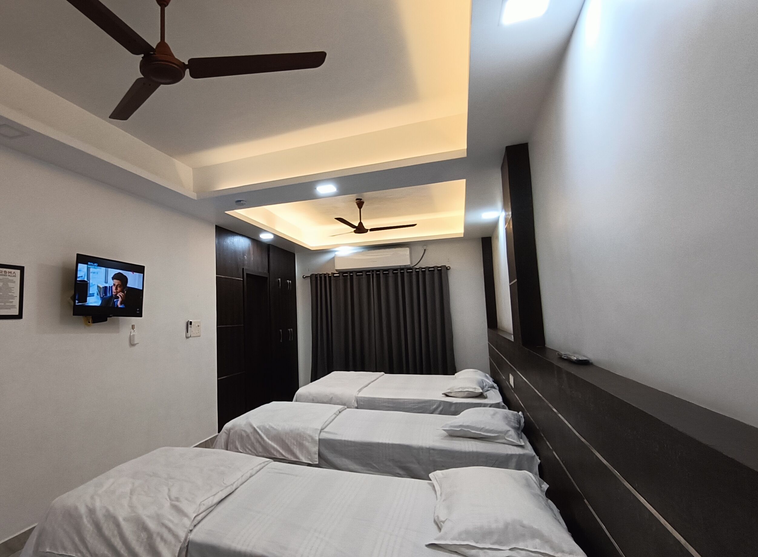 Quarto de hotel com três camas de solteiro, ventilador de teto, televisão fixada na parede e um espelho grande.