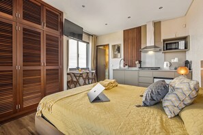 A hotel room with a bed, a laptop, a desk with a chair, a kitchen area, and a sliding door closet.