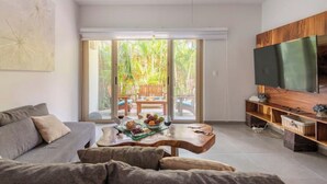 A modern living room with a sofa, a coffee table with fruits and wine, a TV on a wooden stand, and a sliding glass door leading to a patio with a wooden table and chairs.