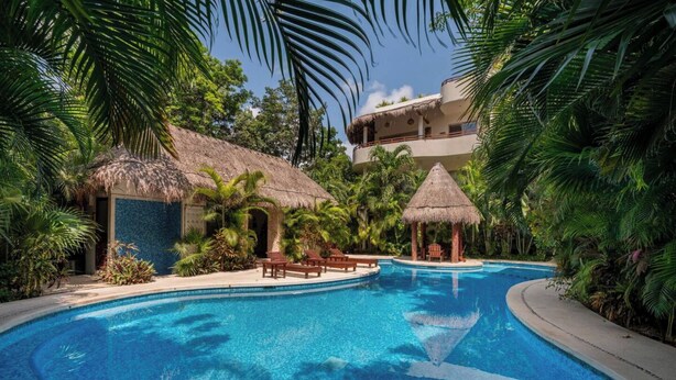 A swimming pool surrounded by palm trees and thatched-roof structures, with a multi-level building in the background.
