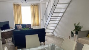 Un soggiorno moderno con un tavolo da pranzo in vetro, sedie bianche, un divano scuro e una scala che conduce a un piano superiore.