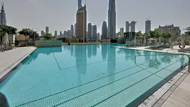 Une piscine à l’eau d’un bleu cristallin, entourée d’une terrasse carrelée. Au fond, on aperçoit plusieurs immeubles de grande hauteur, dont un très grand gratte-ciel.