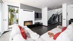 A modern living room with a white sofa, a flat-screen TV, and a fireplace. There is a staircase leading to an upper level. - Villa Clara do Sol - Four Bedroom Villa, Sleeps 8 (Estoi)