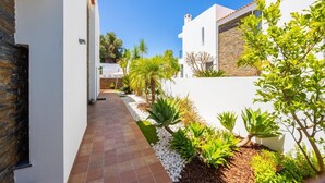A narrow, paved pathway flanked by white walls and green plants, leading to a modern building with a stone facade. - Villa Clara do Sol - Four Bedroom Villa, Sleeps 8 (Estoi)