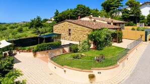 A stone house with a tiled roof, surrounded by a fenced garden and a paved area.