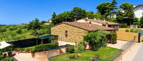 A stone house with a tiled roof, surrounded by a fenced garden and a paved area.