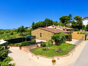 Una casa de piedra con techo de tejas, rodeada por un jardín vallado y una zona pavimentada.