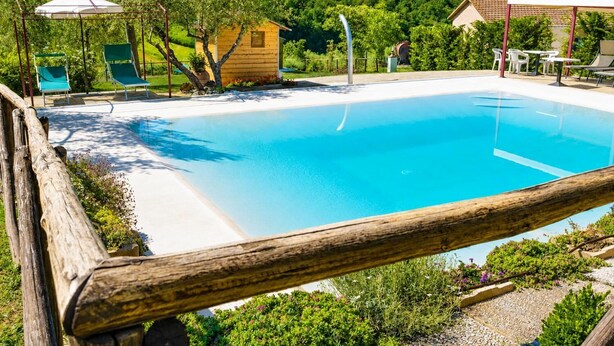 A clear blue pool with a wooden deck and lounge chairs, surrounded by greenery and a small wooden structure.