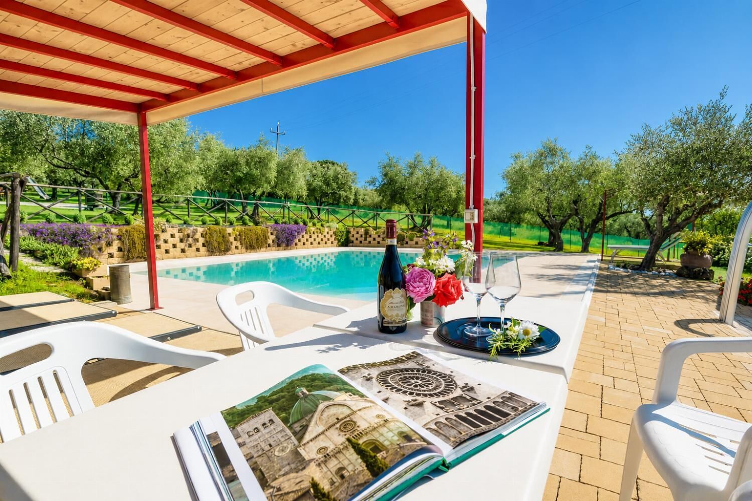 An outdoor patio with a pool, a table with a bottle of wine, flowers, and a book.
