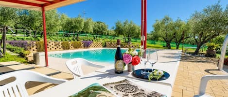 An outdoor patio with a pool, a table with a bottle of wine, flowers, and a book.