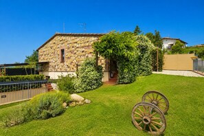 Une maison en pierre avec un toit de pierre, un jardin agrémenté d’une charrette en bois et un ciel d’un bleu limpide.