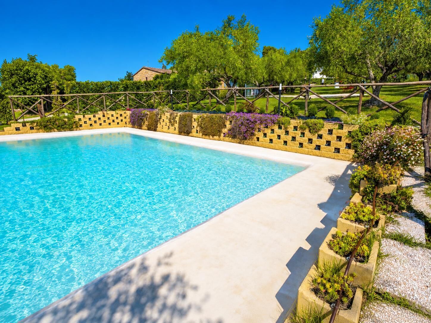 Une piscine rectangulaire à l’eau d’un bleu limpide, entourée d’un petit muret en pierre et d’une végétation luxuriante.