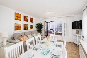 A modern living room with a dining area, white furniture, artwork on the walls, and a view through a glass door.