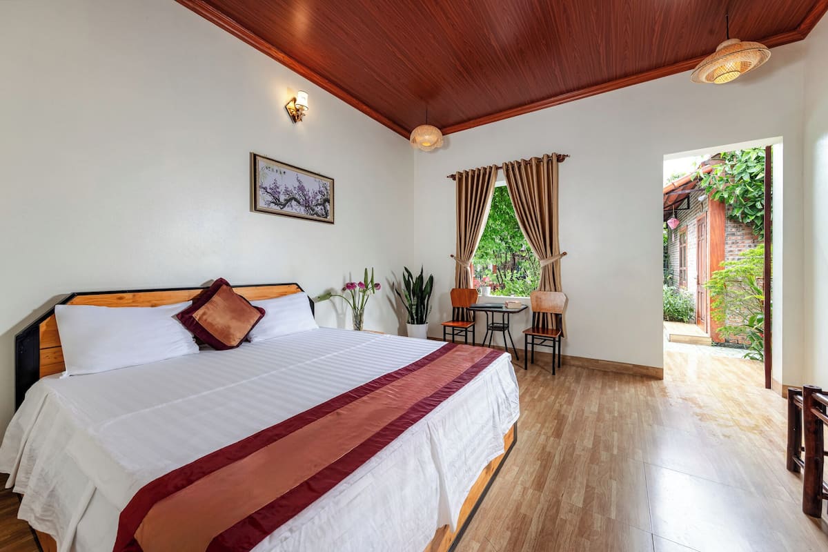 A bedroom with a wooden ceiling, a large bed with a red and white bedspread, a small table with chairs, and a view of greenery outside.