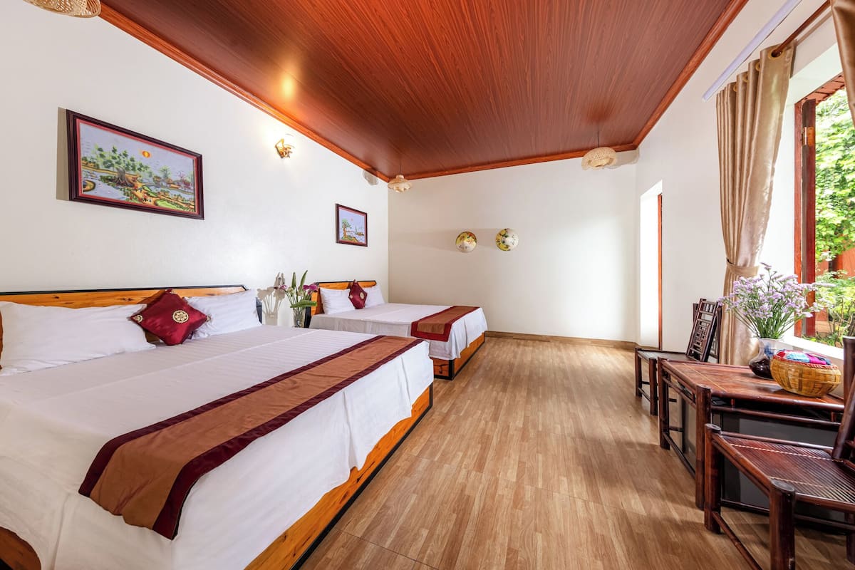 A hotel room with two beds, wooden flooring, a dining table with chairs, and a window with curtains.