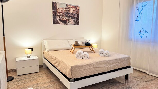 Un lit bien fait, recouvert d’une courtepointe matelassée, une table de chevet avec une lampe, un plateau avec des serviettes et une théière, et un tableau accroché au mur.