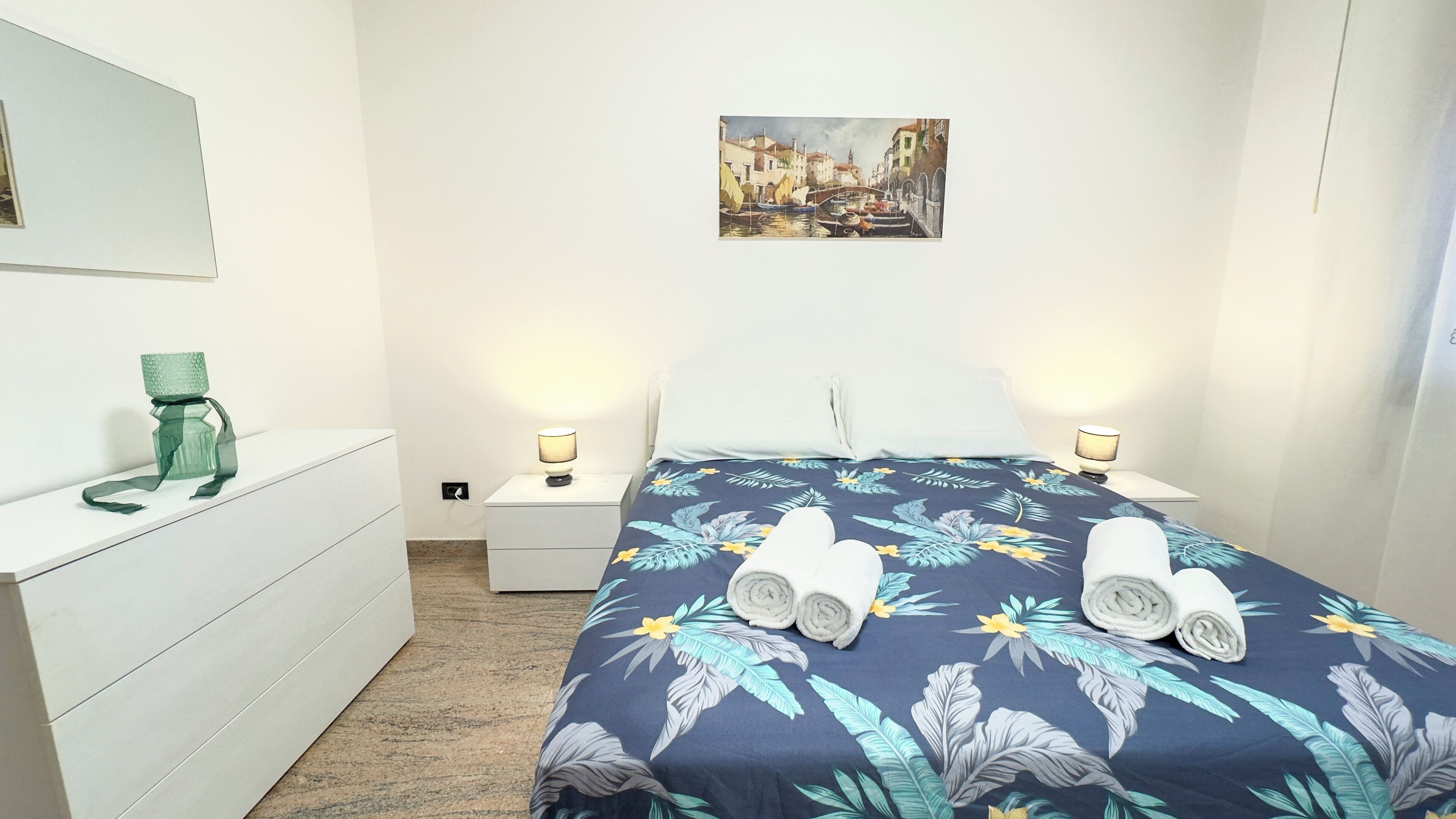 A hotel room with a bed, a nightstand, a dresser, a green bottle, and a painting on the wall.
