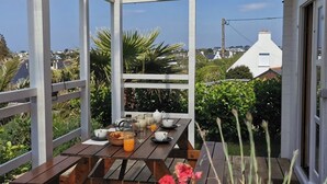 Una terraza de madera con una mesa dispuesta para el desayuno, rodeada de follaje y vistas a casas.