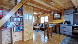 Interior acogedor de una cabaña con una escalera de madera, una sala de estar con sofá y mesa de centro, una cocina con electrodomésticos modernos y una estantería llena de libros.