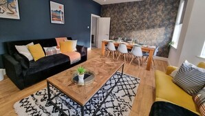 Una sala moderna con un sofá negro, un sofá amarillo y una mesa de centro de madera con una planta. Hay un comedor con una mesa y sillas, y un reloj grande en la pared.