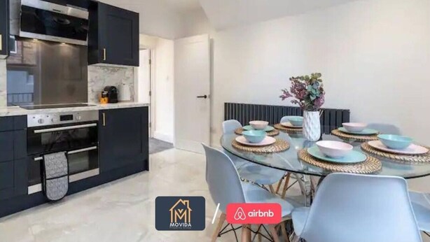 Cocina moderna con armarios oscuros, electrodomésticos de acero inoxidable y una mesa de comedor de vidrio dispuesta con platos y un jarrón de flores.