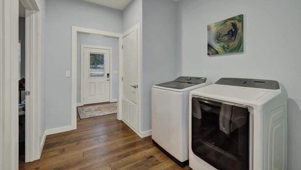 A laundry room with two washing machines and a dryer, wooden flooring, and a door leading to another room.