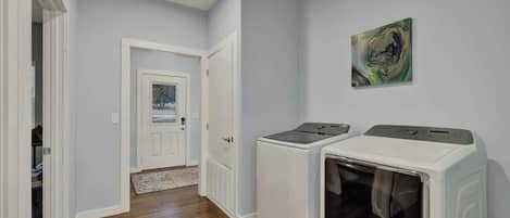 A laundry room with two washing machines and a dryer, wooden flooring, and a door leading to another room.