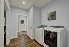 A laundry room with two washing machines and a dryer, wooden flooring, and a door leading to another room.