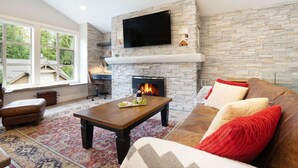 A living room with a stone fireplace, a flat-screen TV mounted above it, a wooden coffee table with a tray of snacks, a leather sofa with red and beige cushions, and a patterned rug.