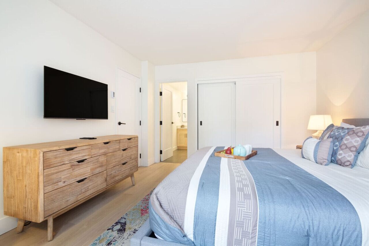 Une chambre à coucher comprenant un lit, un téléviseur fixé au mur, une commode en bois et une porte donnant sur une autre pièce.