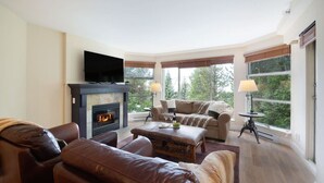A living room with a fireplace, leather sofas, a television, and large windows with views of greenery. - Slopeside ski in/out stylish 2BR/2BA HotTub & Pool (Whistler)