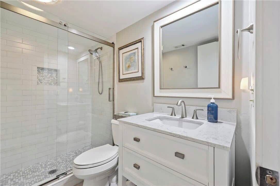 A bathroom with a glass-enclosed shower, a white toilet, a white vanity with a mirror, and a framed picture on the wall.