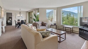 Uma sala de estar moderna com sofá, mesa de centro e televisão, grandes janelas com vista para as montanhas, e um espaço de jantar com cadeiras e mesa.