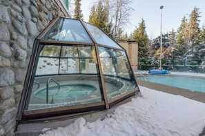 A glass-enclosed hot tub adjacent to a swimming pool, surrounded by snow and trees.