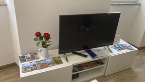 A flat-screen TV on a white entertainment unit with a remote, a vase with red roses, and travel brochures. - 2Curio Court House Charming Lodge (Curio)