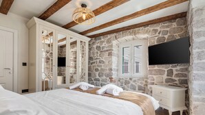 A bedroom with a stone wall, wooden beams, a bed with white linens, a white bedside table, and a mounted TV. - Seaside House - 19th century with Free Parking (Kotor)