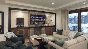 Una sala de estar acogedora con chimenea, televisor montado y vistas a un paisaje nevado a través de amplios ventanales.