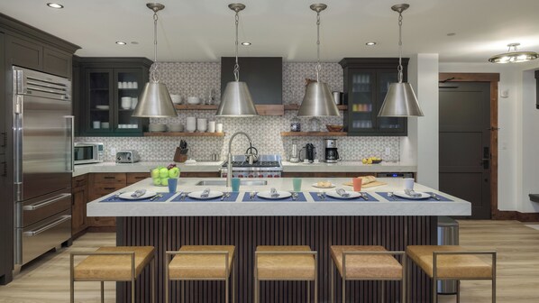 Une cuisine moderne dotée d’un îlot central, d’appareils électroménagers en acier inoxydable, de luminaires suspendus et d’un revêtement mural décoratif.