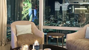 A modern living room with a large window offering a city view, a round black table with candles, and two beige sofas.