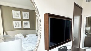 A hotel room with a large mirror, a flat-screen TV, a wooden dresser, and a bed with white linens.