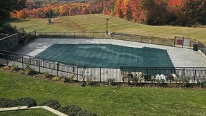 A fenced outdoor area with a green tarp-covered space, surrounded by a well-maintained lawn and trees with autumn foliage.