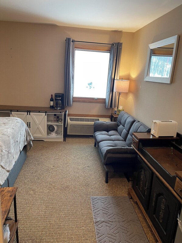A hotel room with a bed, a sofa, a desk with a lamp, a coffee maker, and a window with curtains.