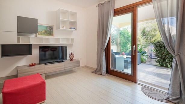 Una sala de estar moderna con un televisor de pantalla plana, un reposapiés rojo y un gran ventanal corredizo que da a un patio con mobiliario exterior.