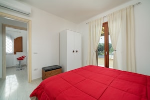 Un dormitorio con colcha roja, un armario, un cofre de mimbre y una silla roja.
