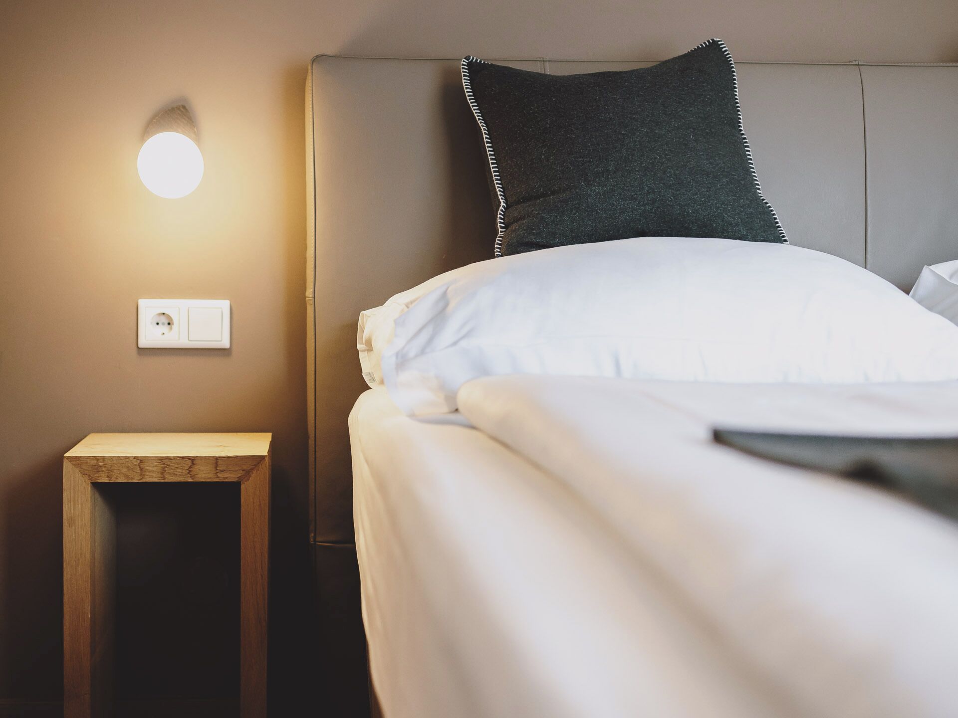 A neatly made bed with white bedding, a small bedside table, a wall socket, and a wall-mounted light fixture.
