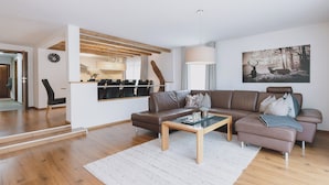 A modern living room with a brown leather sofa, a glass coffee table, and a white rug. There is a dining area with a wooden ceiling and a large window. A painting of deer is on the wall. - Chalet Naila by we rent (Bruck an der Grossglocknerstrasse)